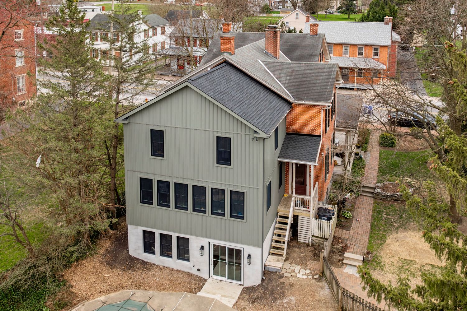 Aerial view of a home with sage green siding and a brick addition amid a wooded neighborhood