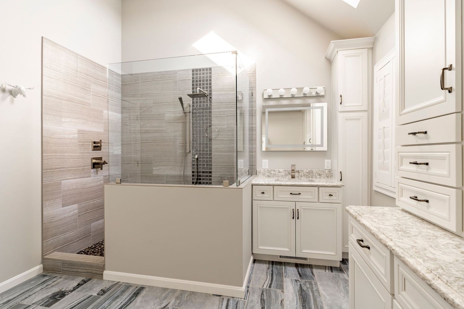 Primary bathroom with a glass shower, gray tile and a white vanity under a lighted mirror