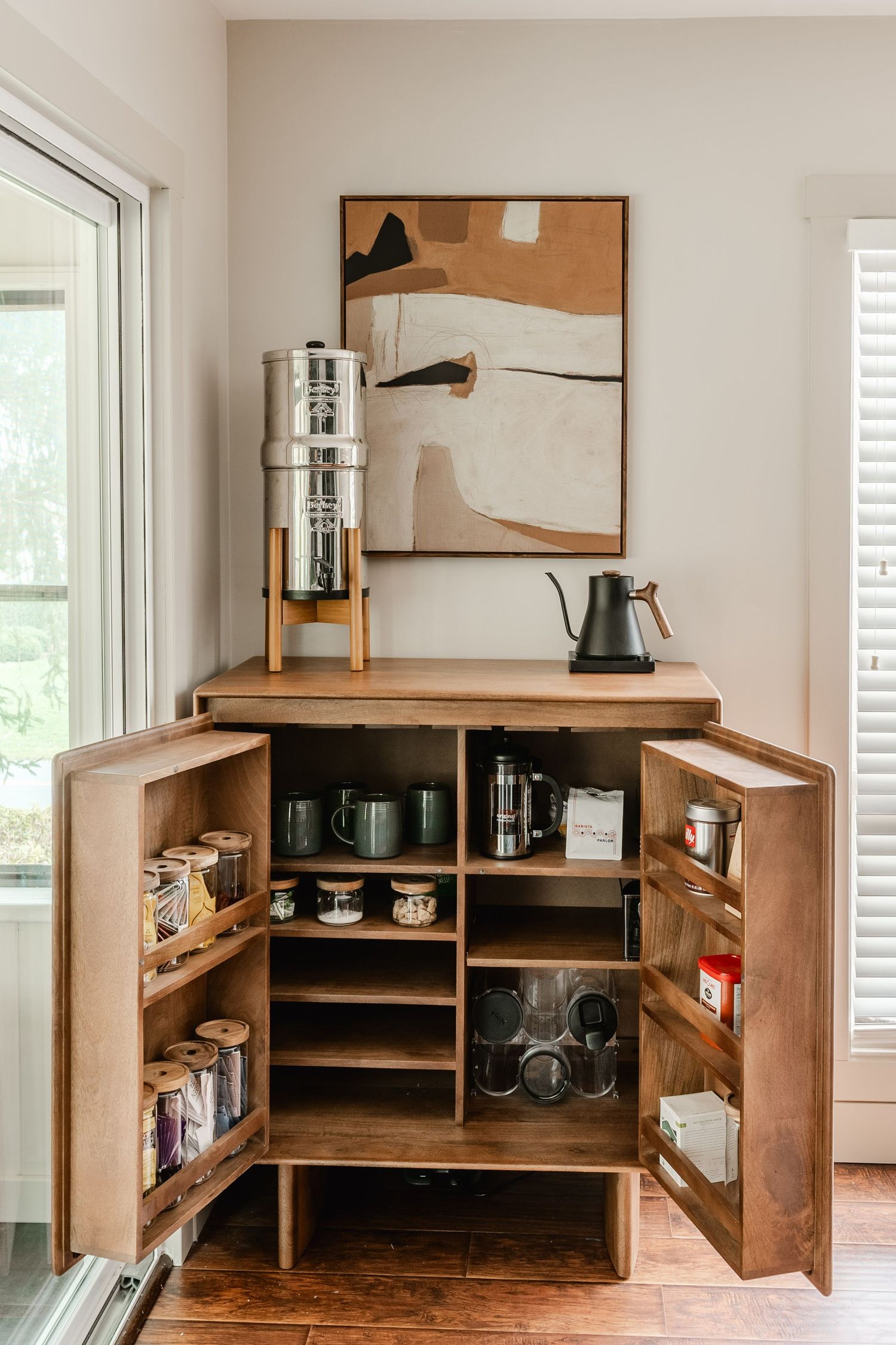 Open wood coffee cabinet with mugs, a French press and a countertop water filter beneath framed art