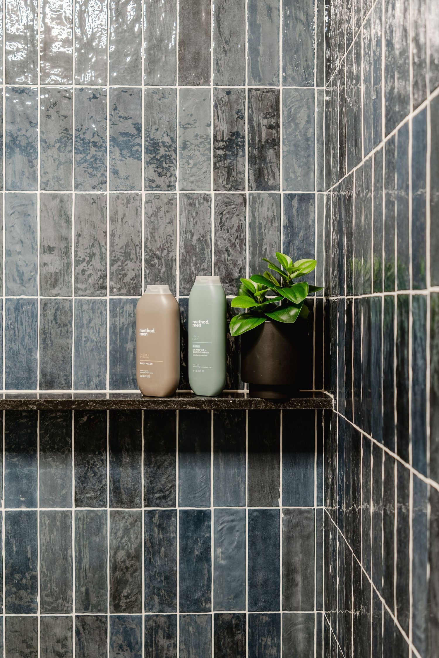 Close-up of a shower niche shelf with body wash bottles and greenery against navy tile