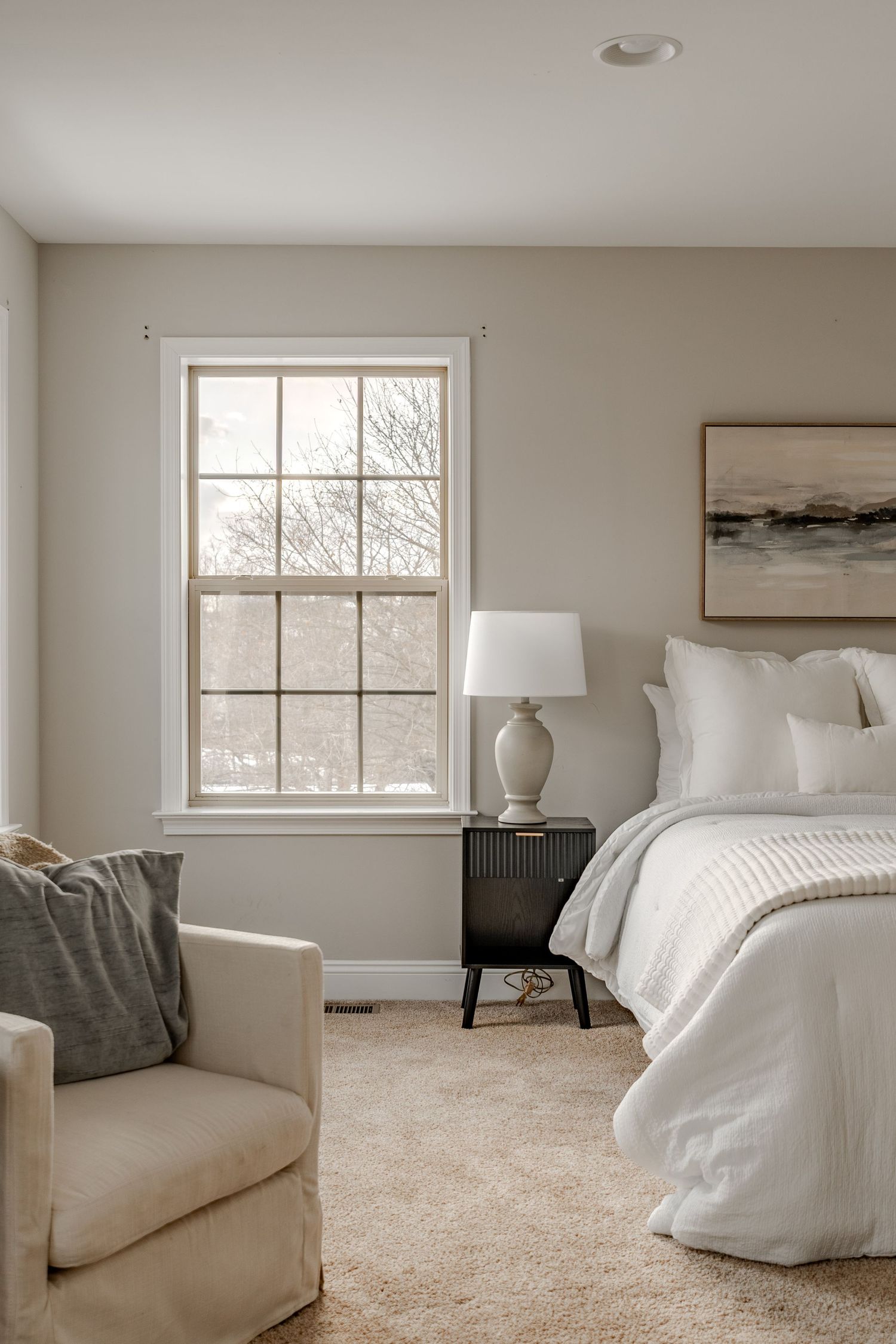 Bedroom with white bedding, a dark wood nightstand, table lamp and framed landscape art