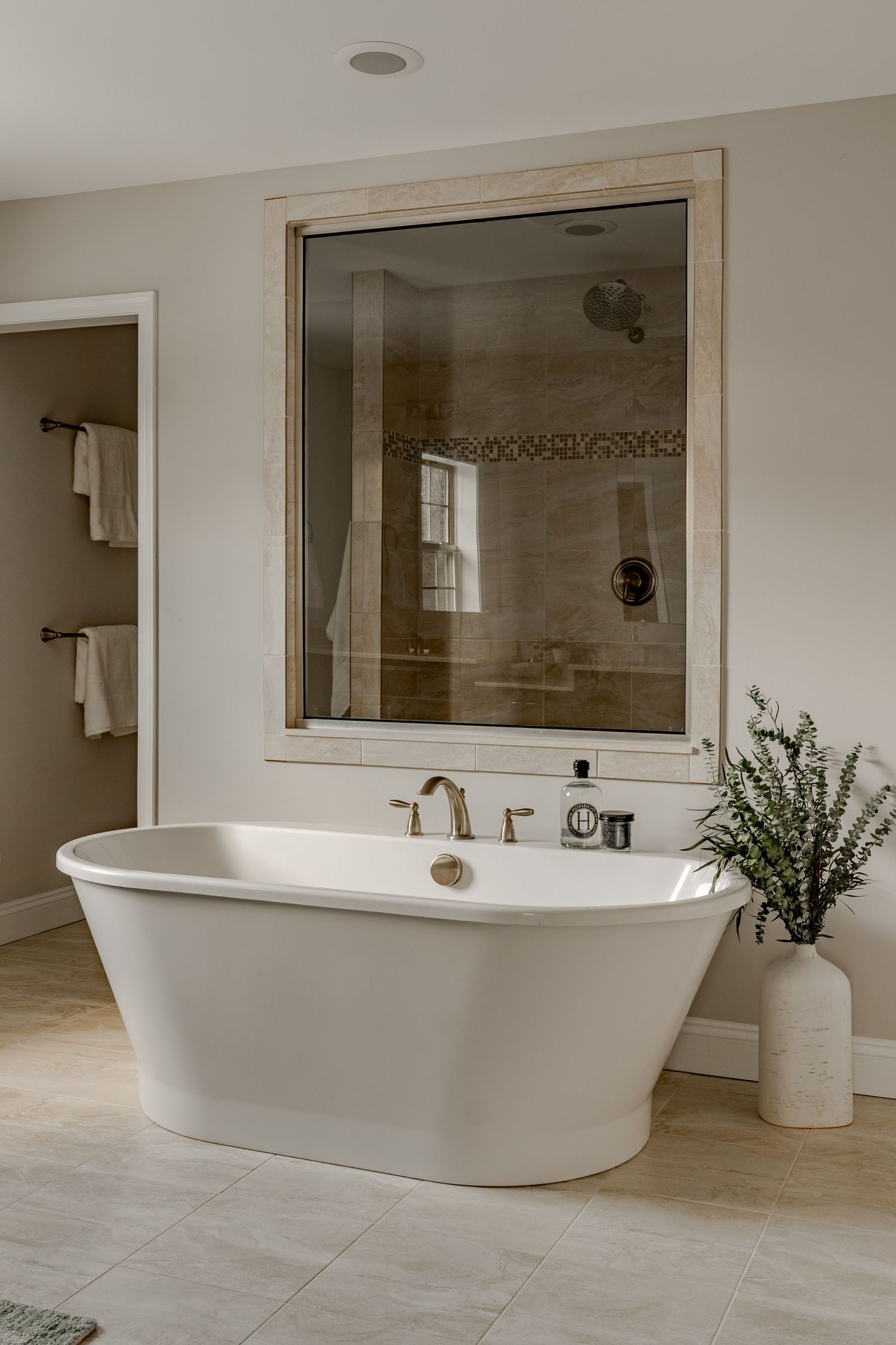 Freestanding tub facing a large mirror-framed window reflecting a tiled walk-in shower