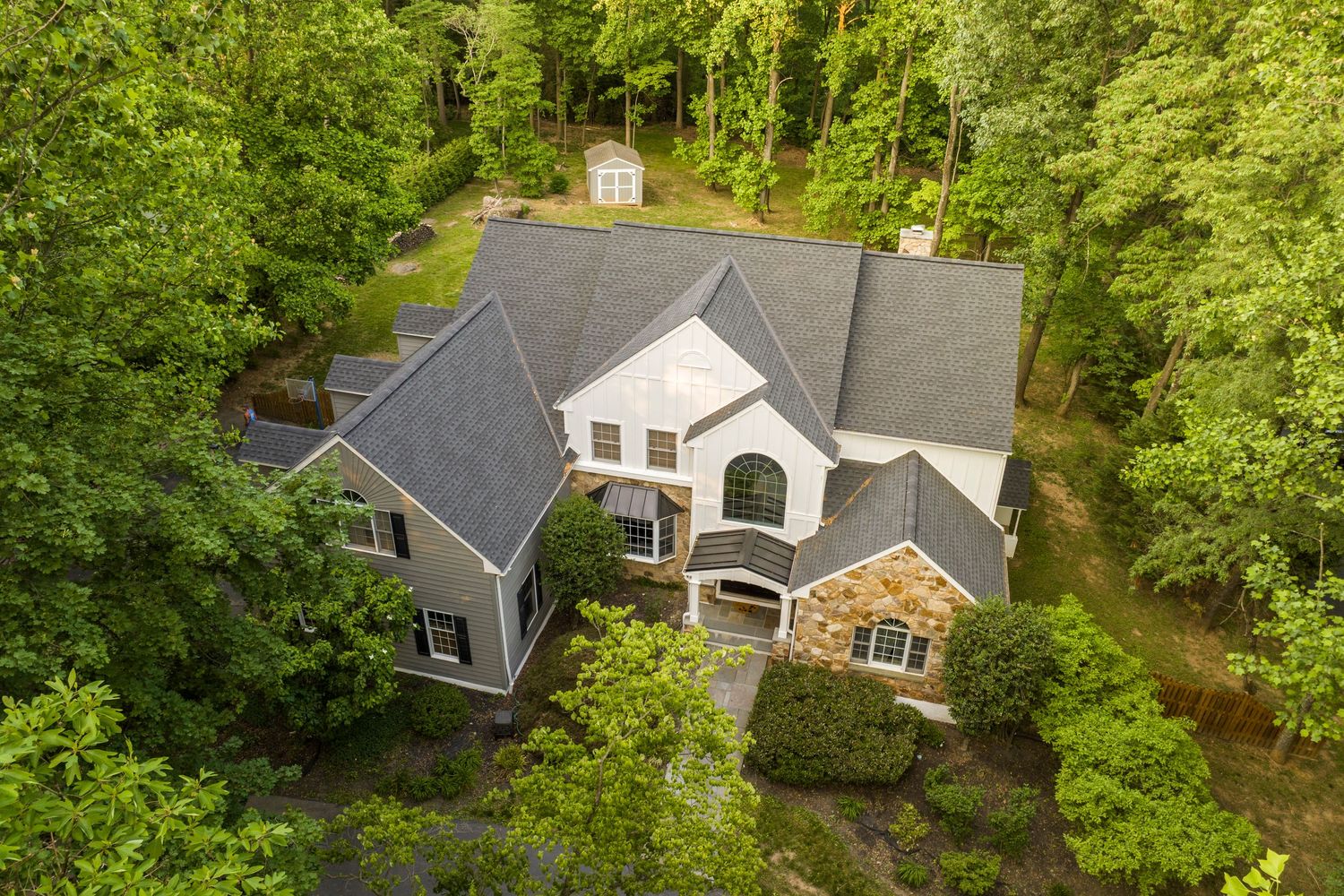 Aerial view of a gray-and-white gabled home tucked into a wooded lot with a storage shed behind it