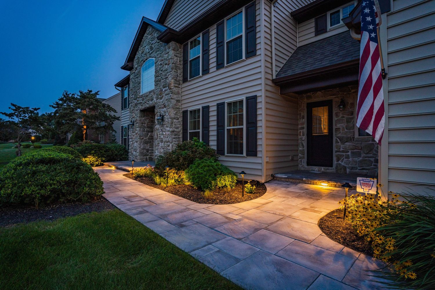 Stone-front home entry at dusk with landscape lighting and an American flag