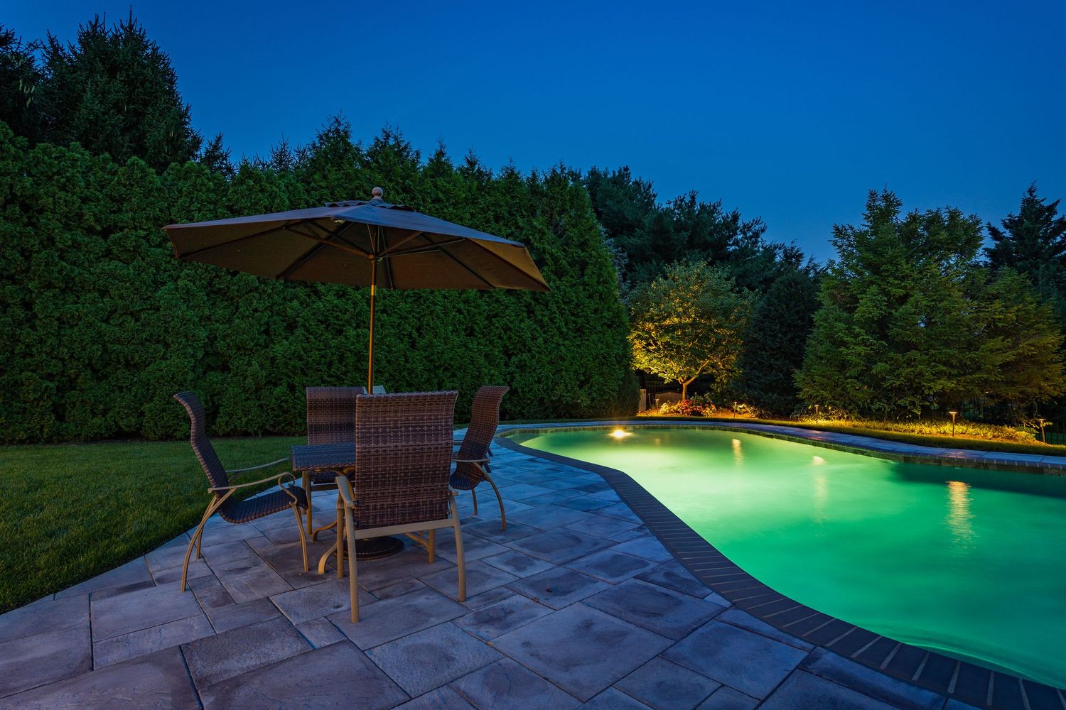 Pool lit green at dusk with a paver patio, wicker dining set and umbrella