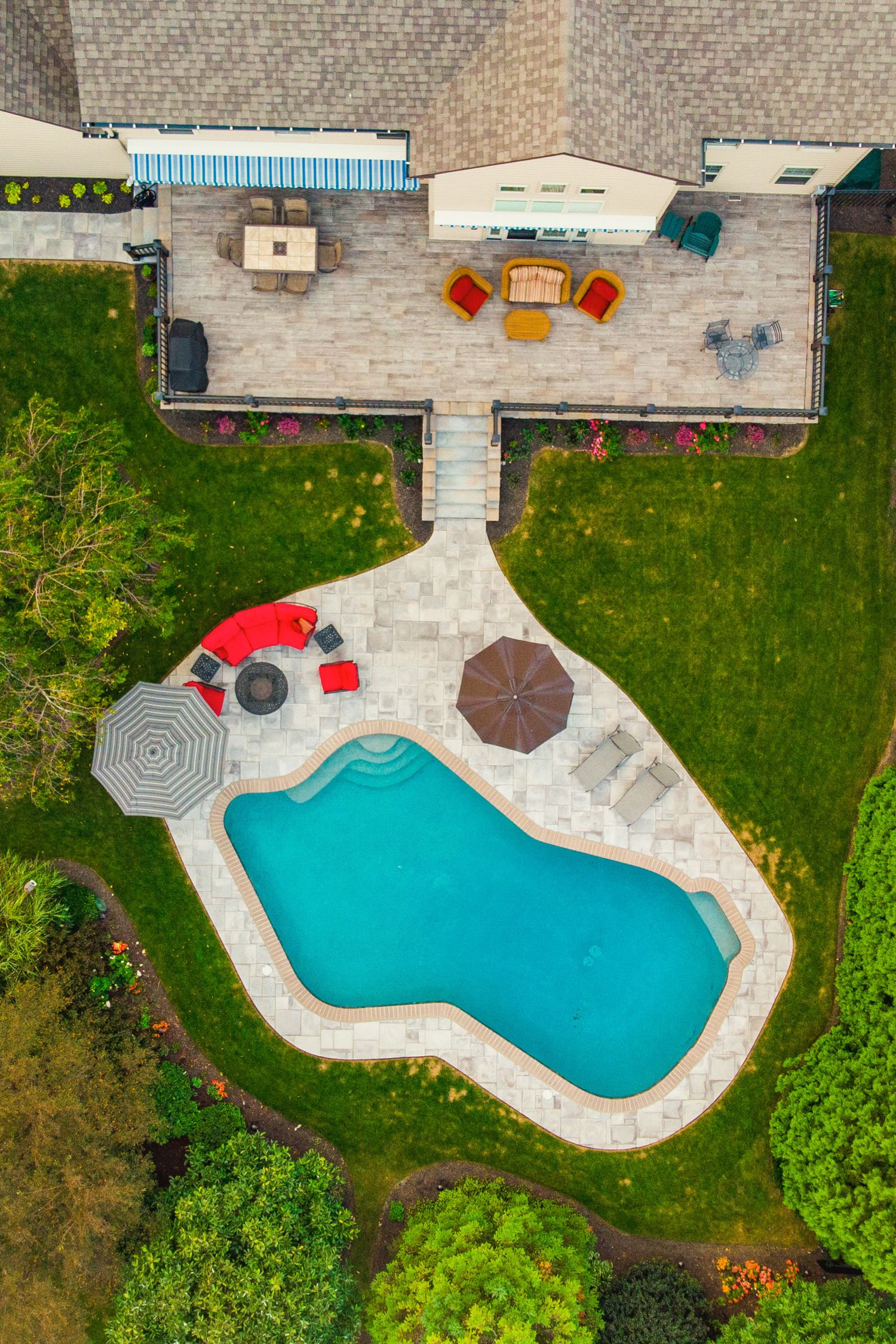 Aerial view of a backyard pool and paver patio with red furniture and market umbrellas