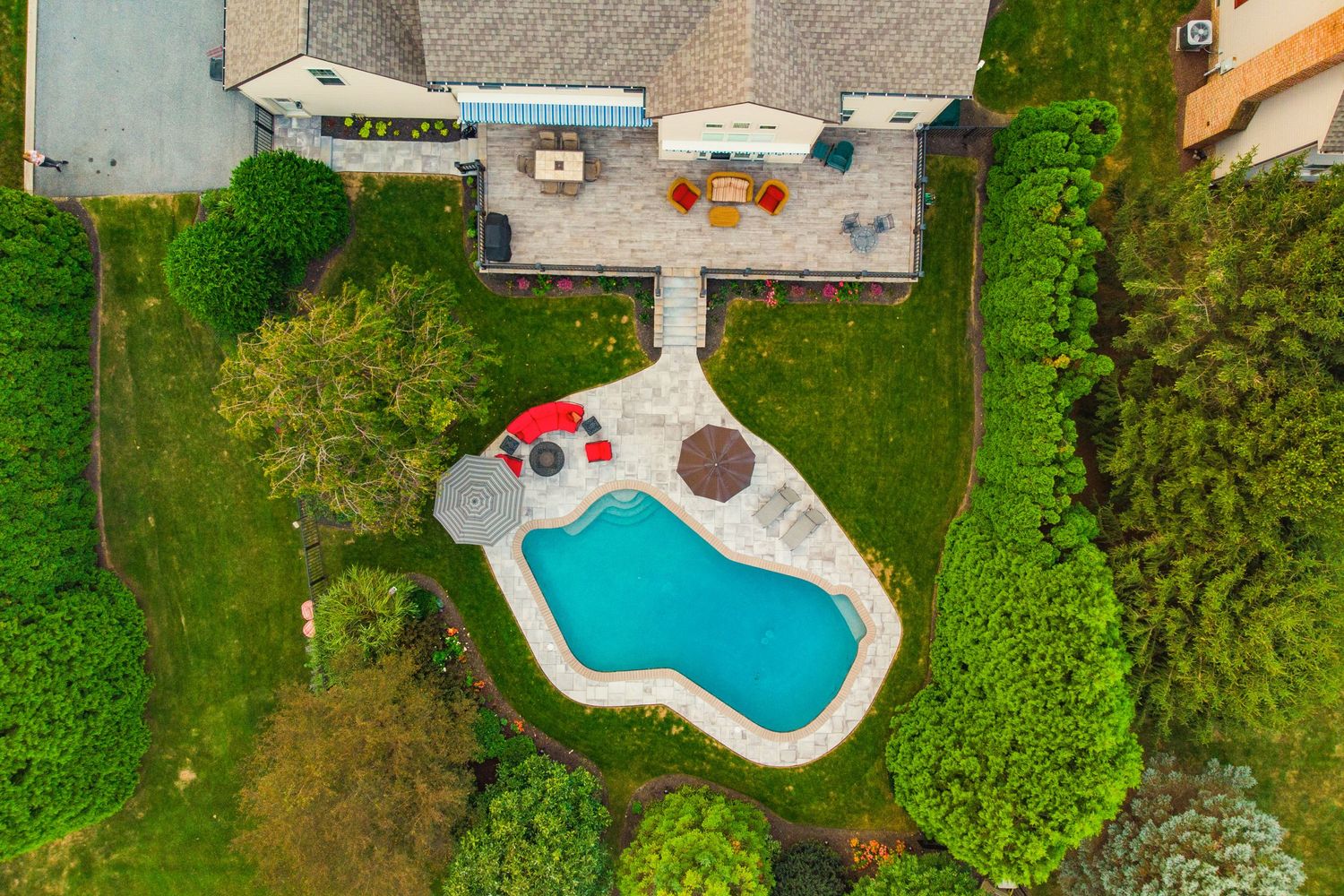 Aerial view over a home's rooftop showing a furnished deck, stone walkway and curved pool below