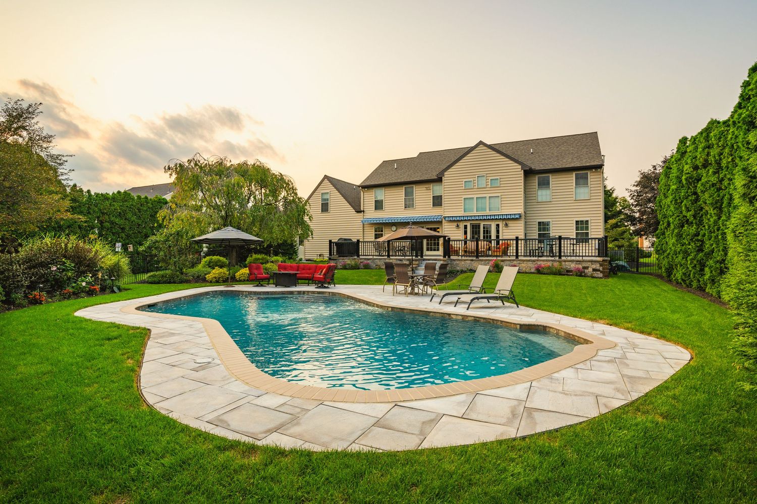 Backyard pool with paver patio, red outdoor furniture and a two-story home at dusk