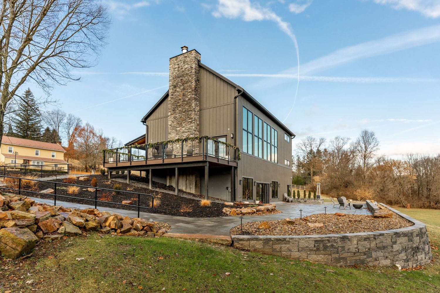 Rear exterior of a farmhouse with stone chimney, elevated deck and curved stone retaining wall