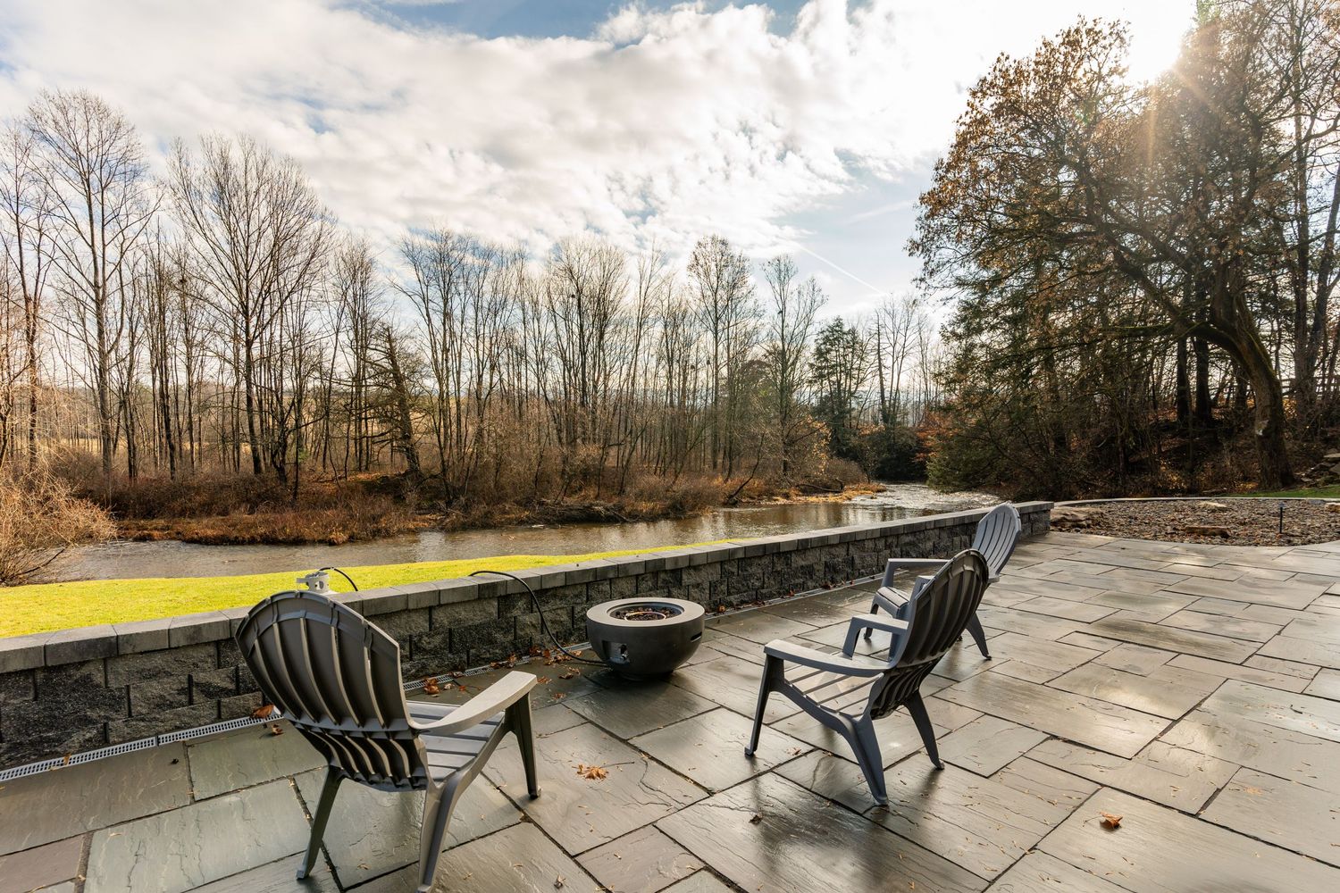 Patio seating around a fire pit beside a stone wall overlooking a wooded creek at sunset
