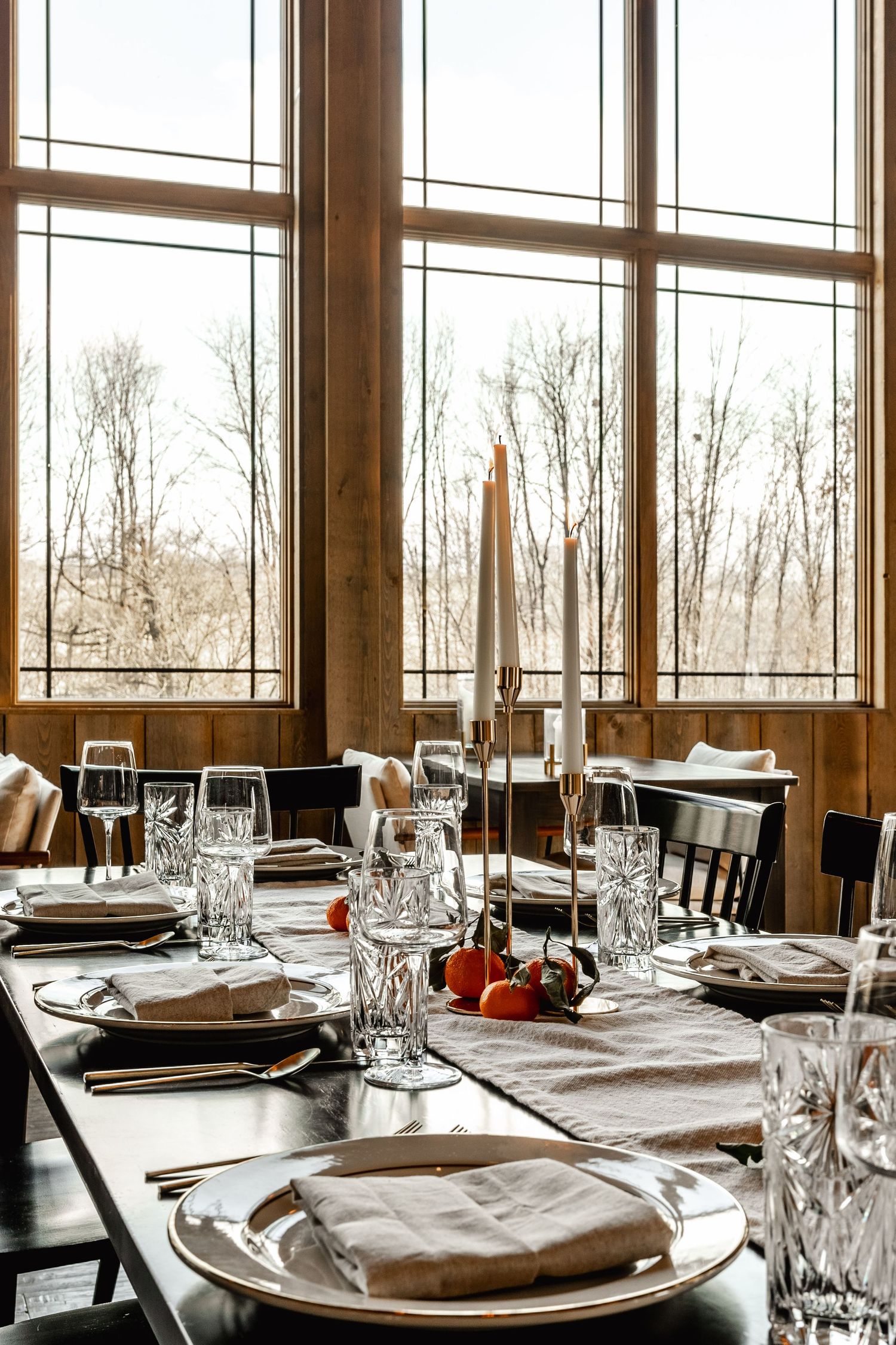 Close-up table setting with taper candles and gold place settings beneath tall wood-framed windows