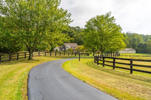 Paved driveway winding between mature trees and a black wood fence toward a farmhouse