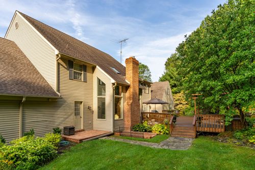 Two-story tan-sided home with a covered deck and pergola surrounded by mature trees