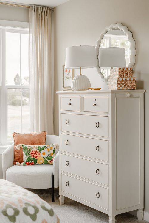 White dresser with a scalloped mirror, ceramic lamp and patterned boxes beside a peach chair