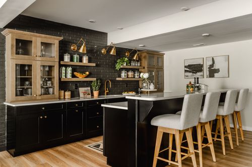 Basement bar with black cabinetry, glass-front hutches, gold sconces and barstool seating