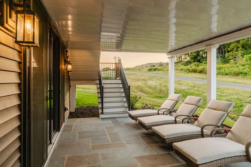 Covered stone patio with wicker chaise lounges beside an exterior staircase at dusk