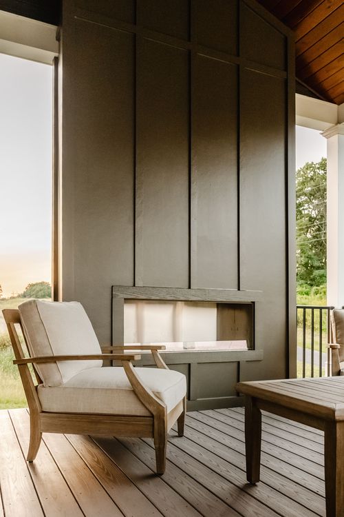 Cushioned wood armchair beside an outdoor linear fireplace on a covered porch