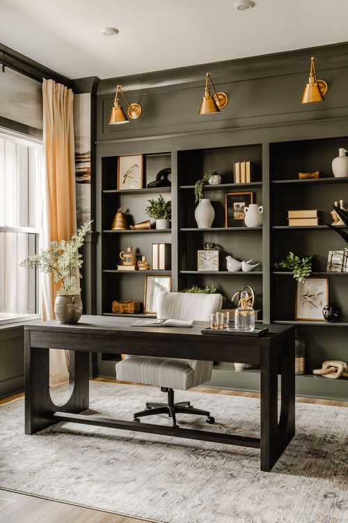 Home office with olive-green built-in shelving, brass wall sconces, and a dark desk