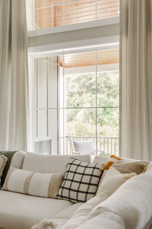 Layered throw pillows on a cream sectional sofa beside floor-to-ceiling windows and a balcony
