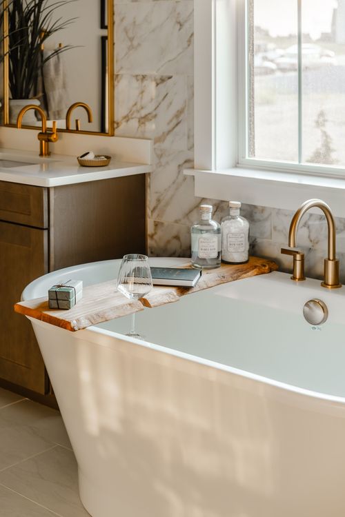 Freestanding soaking tub with a wood tray, wine glass and brass filler in a marble bathroom