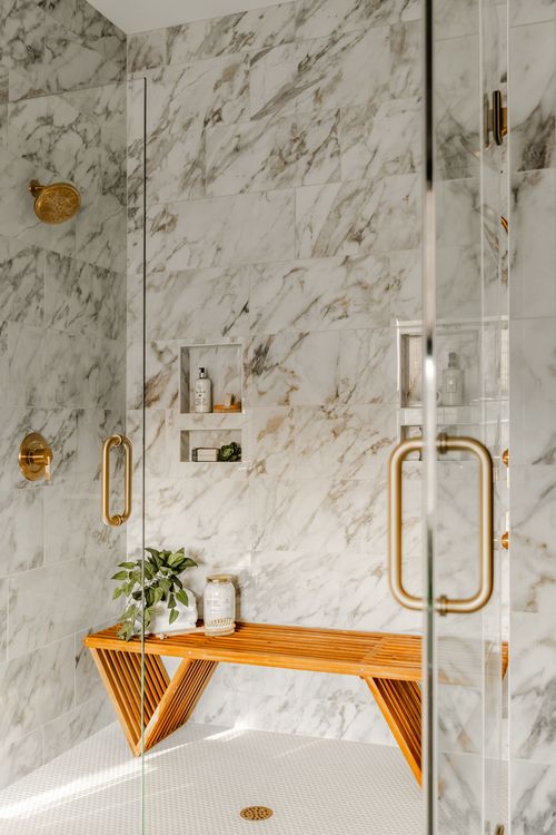 Walk-in shower with marble tile, brass fixtures, glass doors and a teak bench