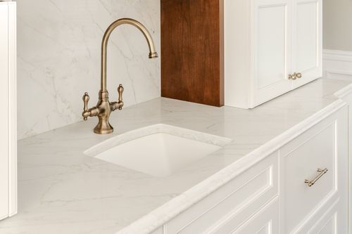 White marble counter with a brass faucet and undermount sink beside cream cabinet drawers