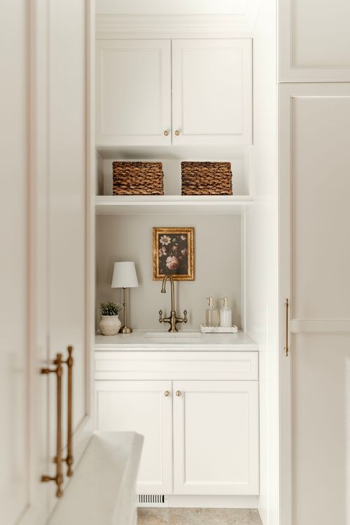White cabinetry flanking a marble sink nook with woven baskets and framed floral art