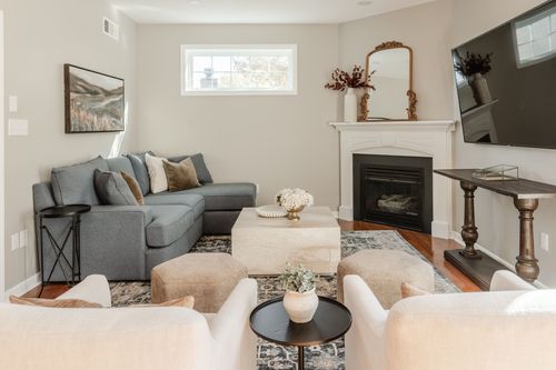 Living room with a blue-gray sectional, cream ottomans, corner fireplace and gold mirror
