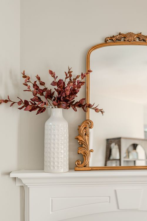 Close-up of a gold ornate mirror and dried florals in a white vase on a mantel