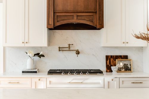 Gas cooktop beneath a carved wood range hood with a brass pot filler and marble backsplash