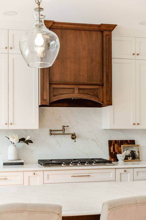 Wood range hood with a glass pendant light beside a gas cooktop and marble backsplash