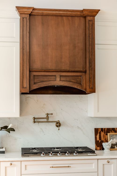 Dark wood range hood with brass pot filler over a marble slab backsplash in a white kitchen