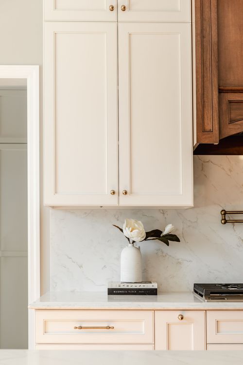 Cream cabinetry with brass knobs and a marble backsplash beside a vase of white magnolia