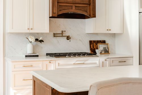 Dark wood range hood between white cabinets with a marble backsplash and brass pot filler