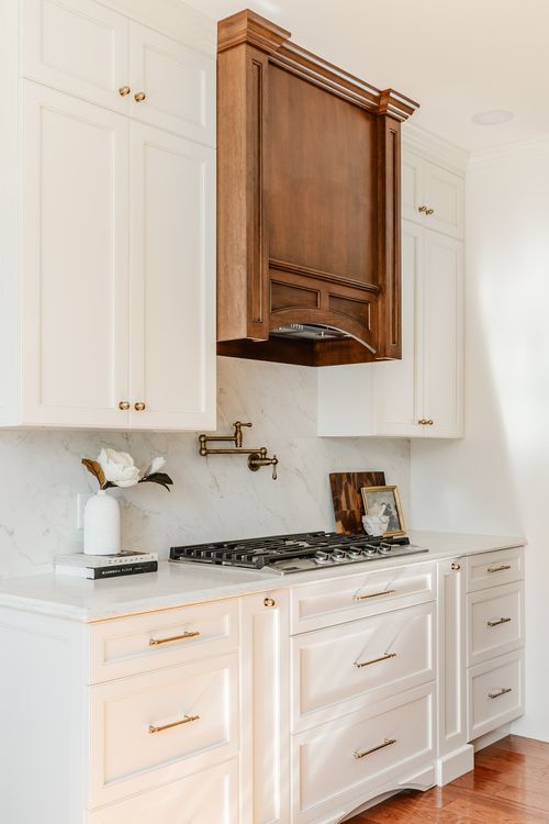 Stainless gas cooktop beneath a dark wood range hood with white cabinetry and a marble backsplash