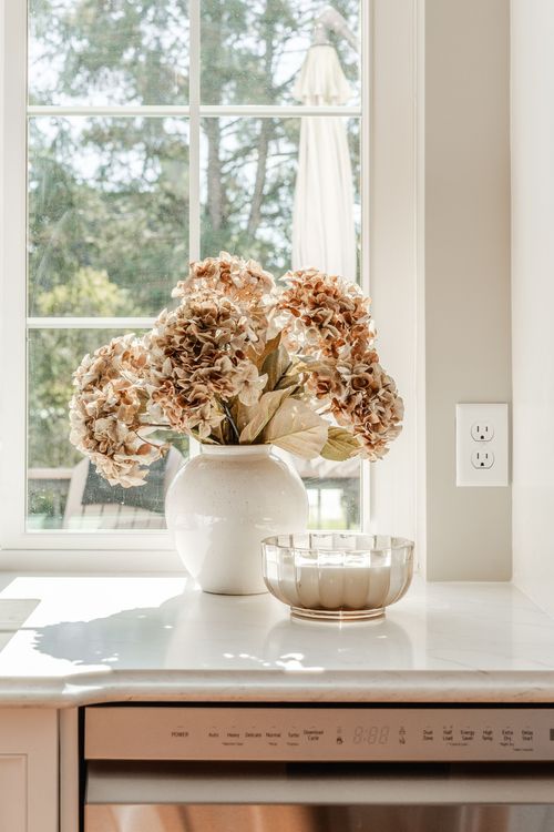 Dried hydrangea arrangement and a glass candle bowl on a marble kitchen counter by a window