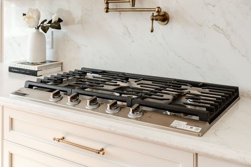 Close-up of a stainless gas cooktop with a brass pot filler and marble backsplash