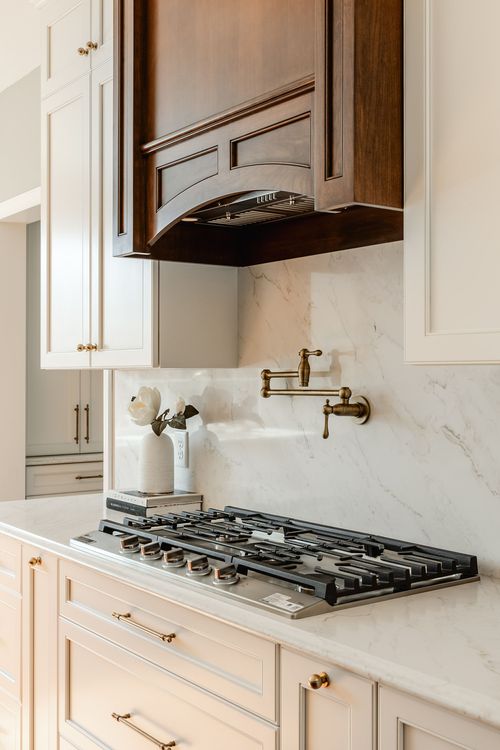 Arched wood range hood over a gas cooktop with brass pot filler and white cabinetry