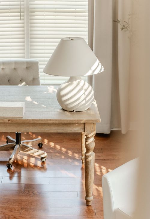 Close-up of a swirled white table lamp on a wood desk beside sheer curtains