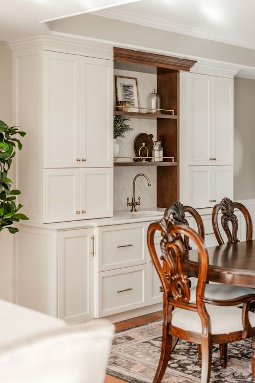 White built-in cabinetry surrounding a wood bar niche with brass faucet and carved dining chairs