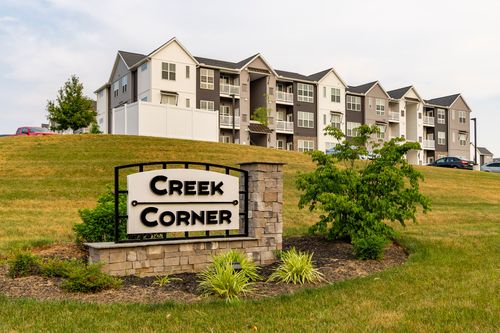 Ground-level view of the Creek Corner sign framed by shrubs with an apartment building behind