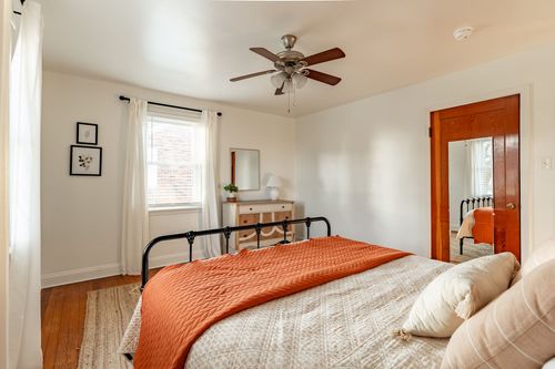 Bedroom with a black iron bed, orange throw, wood dresser and corner windows with sheer curtains