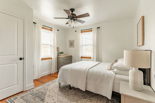 Corner bedroom with a gray dresser, ceiling fan and white bedding beside double windows