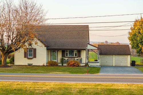 Front exterior of a cream cape cod home with a covered porch and attached garage under fall trees