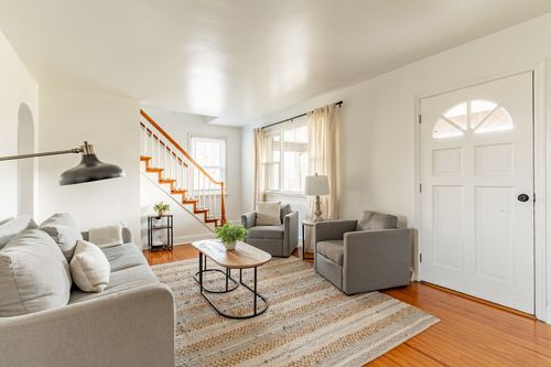 Open living room with gray seating, a staircase with orange-painted railing and a glass-paned front door