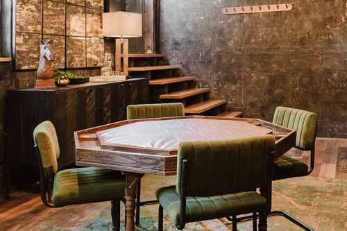 Octagonal poker table with green velvet chairs beside a dark accent wall and staircase