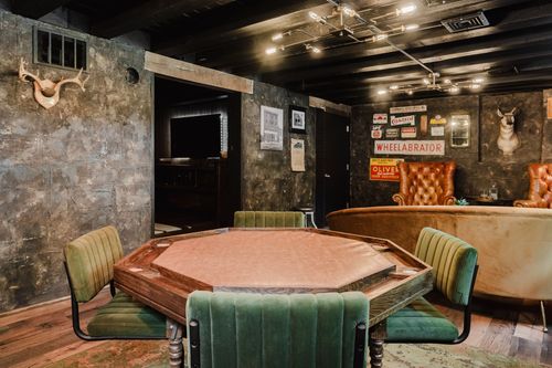 Game room with a poker table and green chairs facing leather sofas under mounted antlers