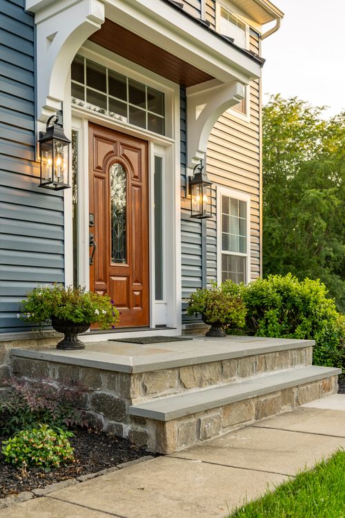 Front entry with a white arched overhang, wood door and stone steps with planted urns