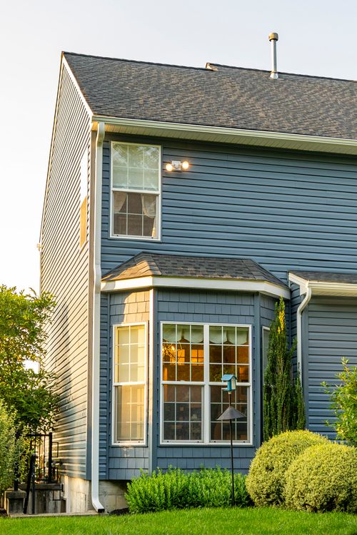 Bay window with white trim on a blue two-story home, lit warmly at dusk