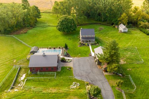 Aerial view of a stone farmhouse with a pool, red barn and long driveway amid green fields